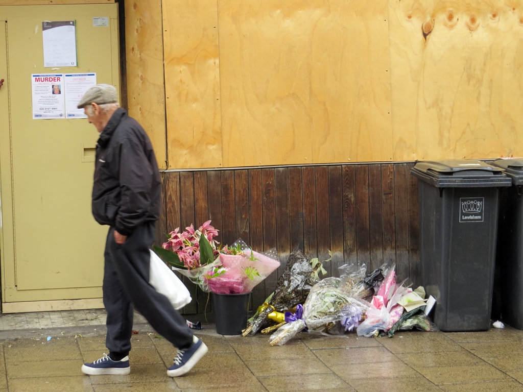 Old man with a cap walks past flowers outside boarded up cafe  on Deptford High Street left in tribute to  arson attack victim