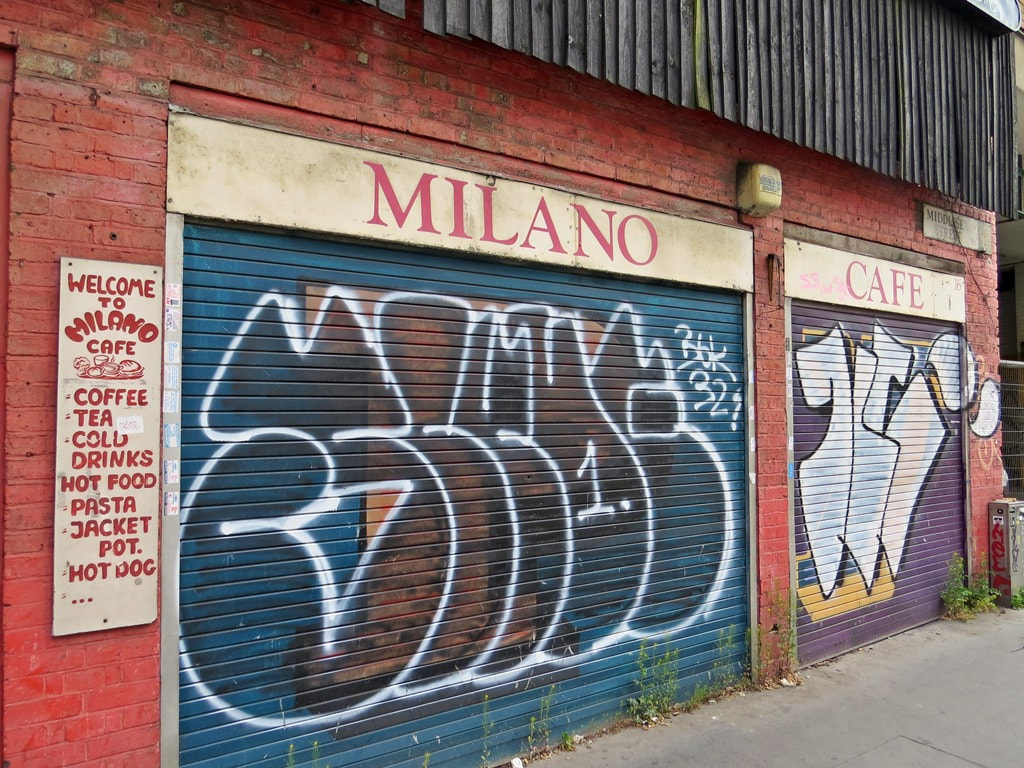 Graffiti covered shutters for derelict Milano Cafe and old sign advertising tea, coffee, hot food, pasta, jacket potato and hot dogs